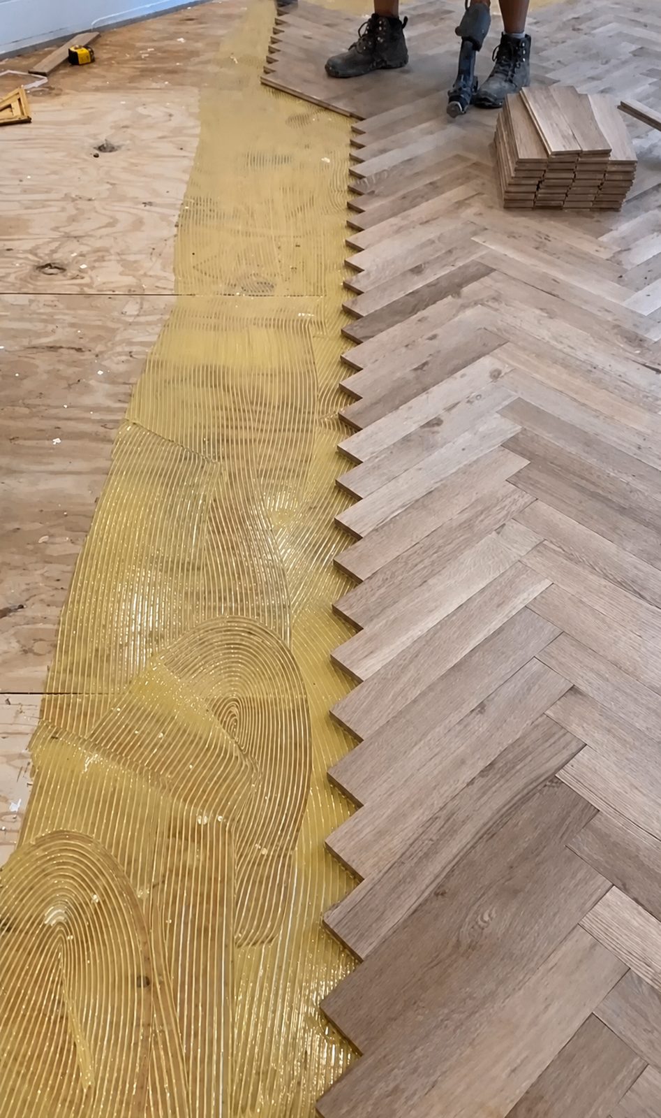 Herringbone hardwood planks being set into fresh adhesive during a new floor installation.